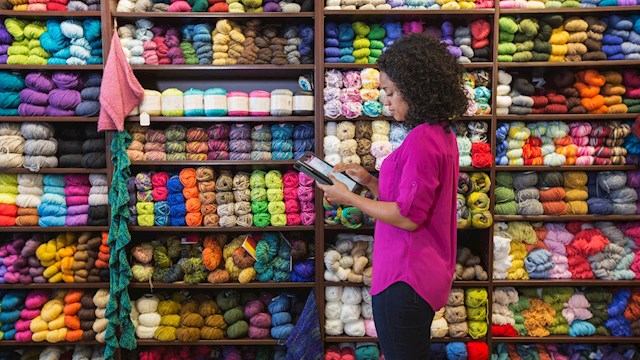 Woman managing inventory in a colorful yarn store using a digital device, representing small business retail logistics powered by smart fulfillment solutions.